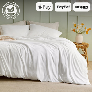 Luxurious white bed with soft, draped sheets in a cozy bedroom setting. The bed is adorned with pillows and a light beige throw blanket. A vase with fresh flowers sits on a wicker side table, adding a touch of nature to the room. The image includes logos for Apple Pay, PayPal, and Shop Pay, highlighting convenient payment options. The '100% Natural Organic Product' badge emphasizes the eco-friendly aspect of the bedding