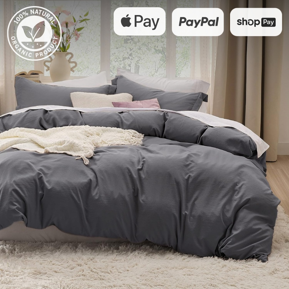 Charcoal bedding with white throw, pink pillow in a bright room with large window, vase, and cozy decor, eco-friendly logos at top.