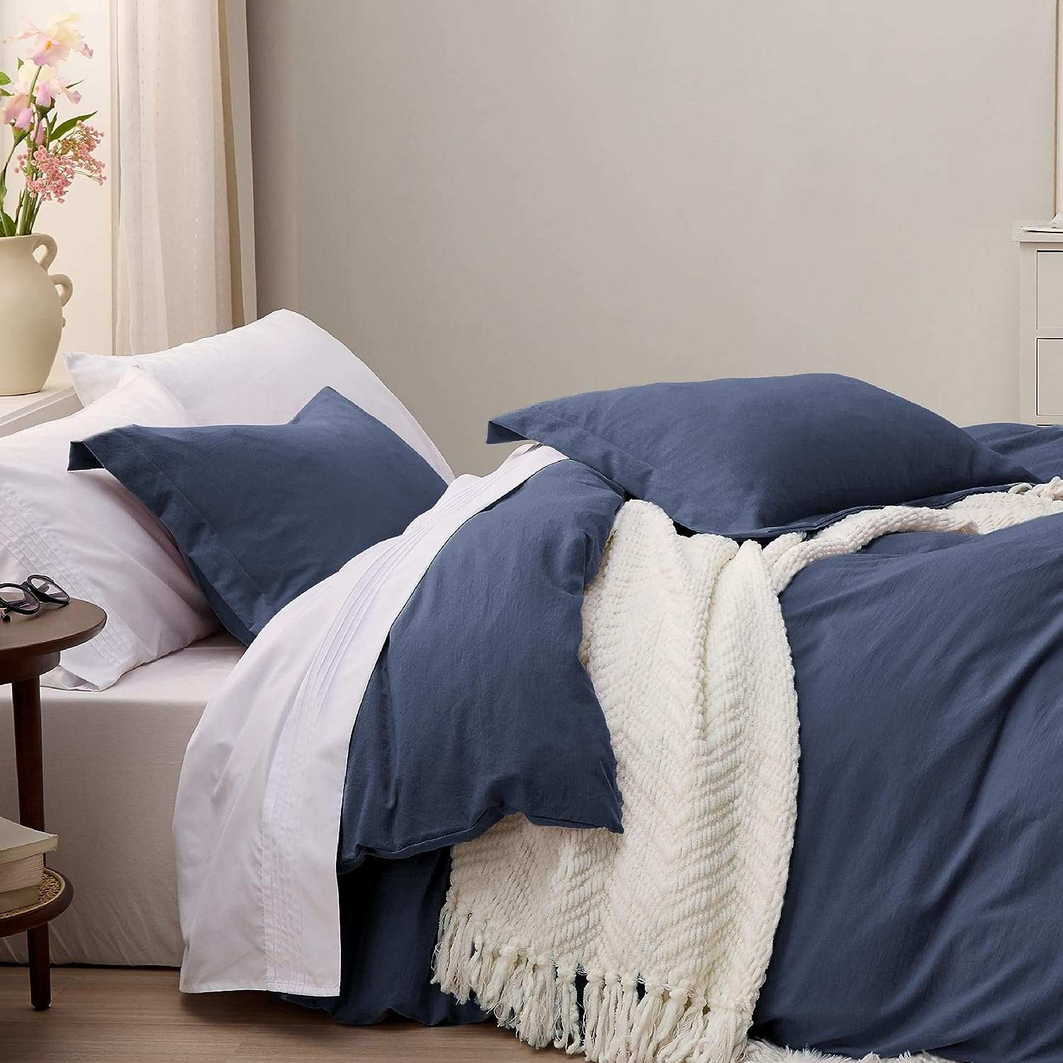 Bed with navy blue bedding and white sheets, adorned with a white textured blanket with fringe. The bed is against a light gray wall, with a bedside table holding a pair of glasses and books. A vase with pink flowers is visible in the background, adding a touch of color and freshness to the room