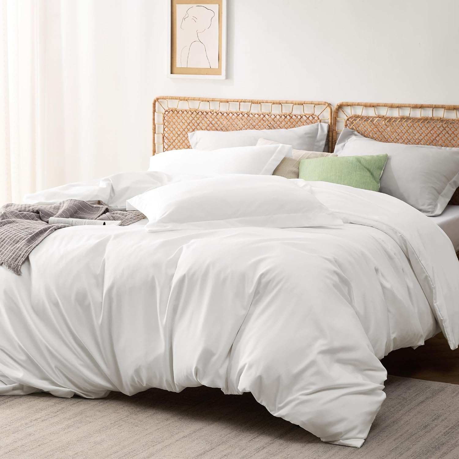 Luxurious white bed with plush bedding and pillows, set in a serene bedroom with light decor. The bed features a rattan headboard and a mix of white and green pillows. A gray throw blanket is casually placed on the bed, alongside a magazine. Above the bed, a simple line art portrait hangs on the wall, complementing the minimalist and cozy aesthetic