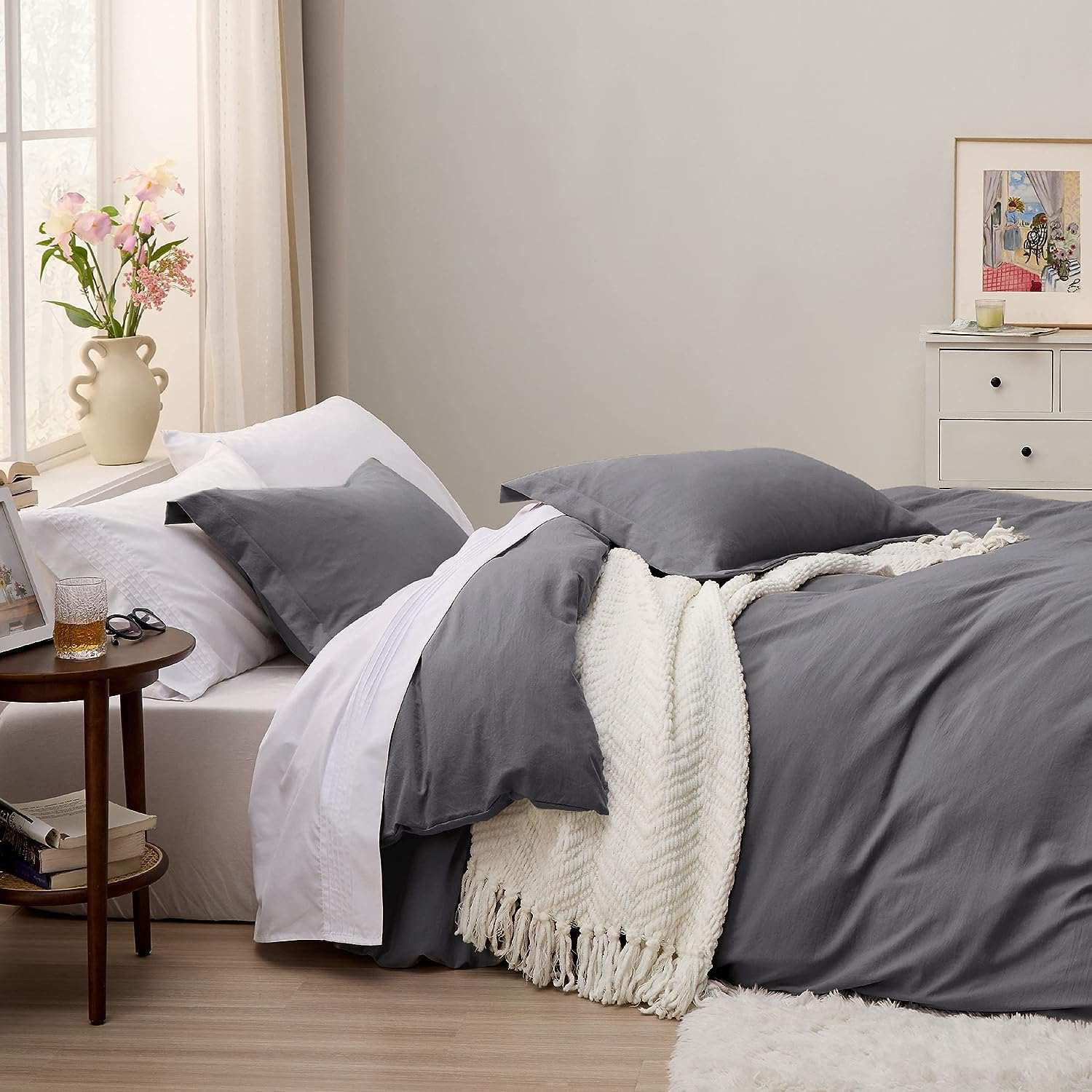 Charcoal bedding with white throw in a bright bedroom, soft lighting, floral vase, and cozy decor create a peaceful atmosphere.