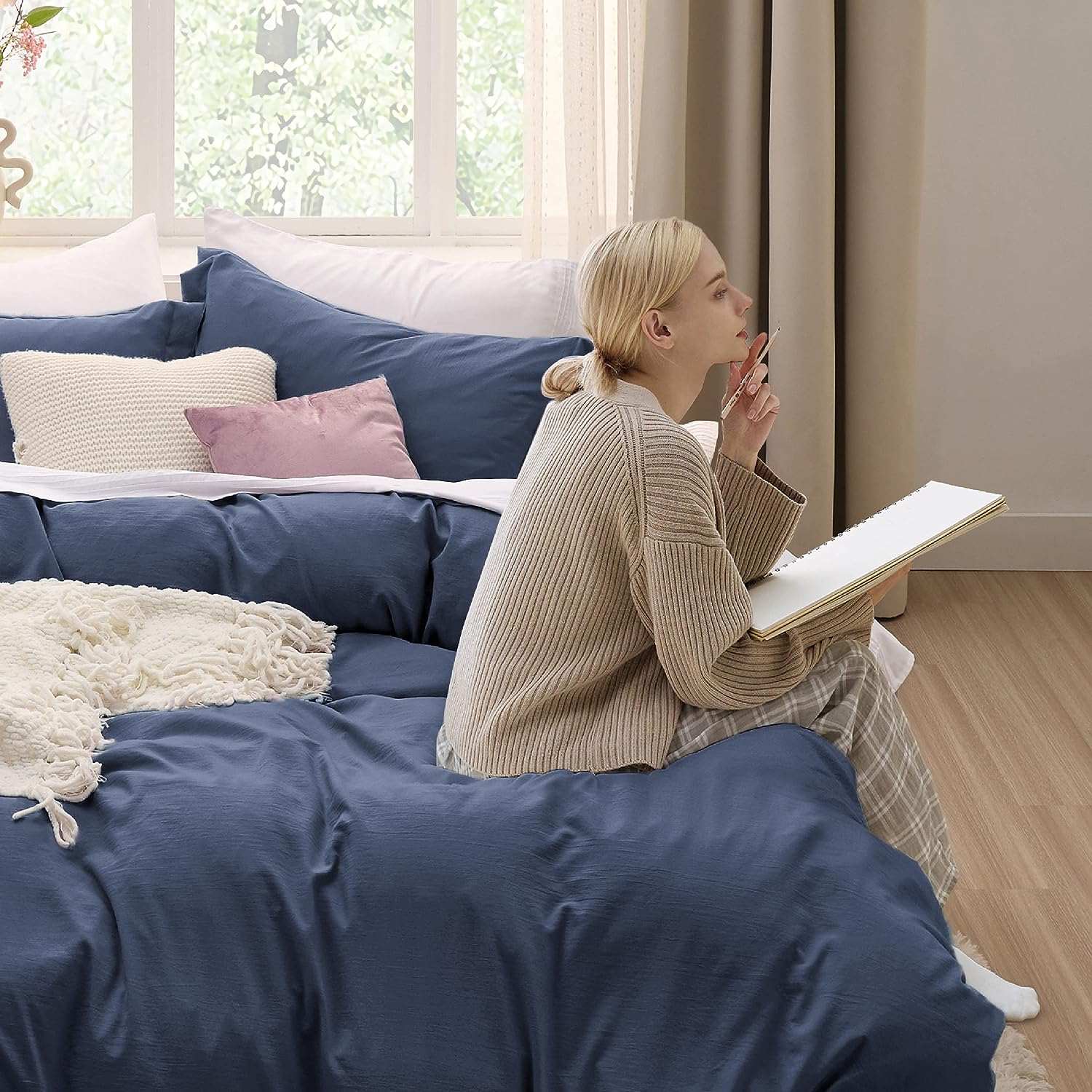 A woman with blonde hair tied in a low bun sits on a bed with navy blue bedding, holding a notebook and pen, looking thoughtfully out of the window. The bed is adorned with white and blush pink pillows, along with a textured white blanket. The background features large windows with sheer curtains, letting in natural light and showcasing a leafy view.