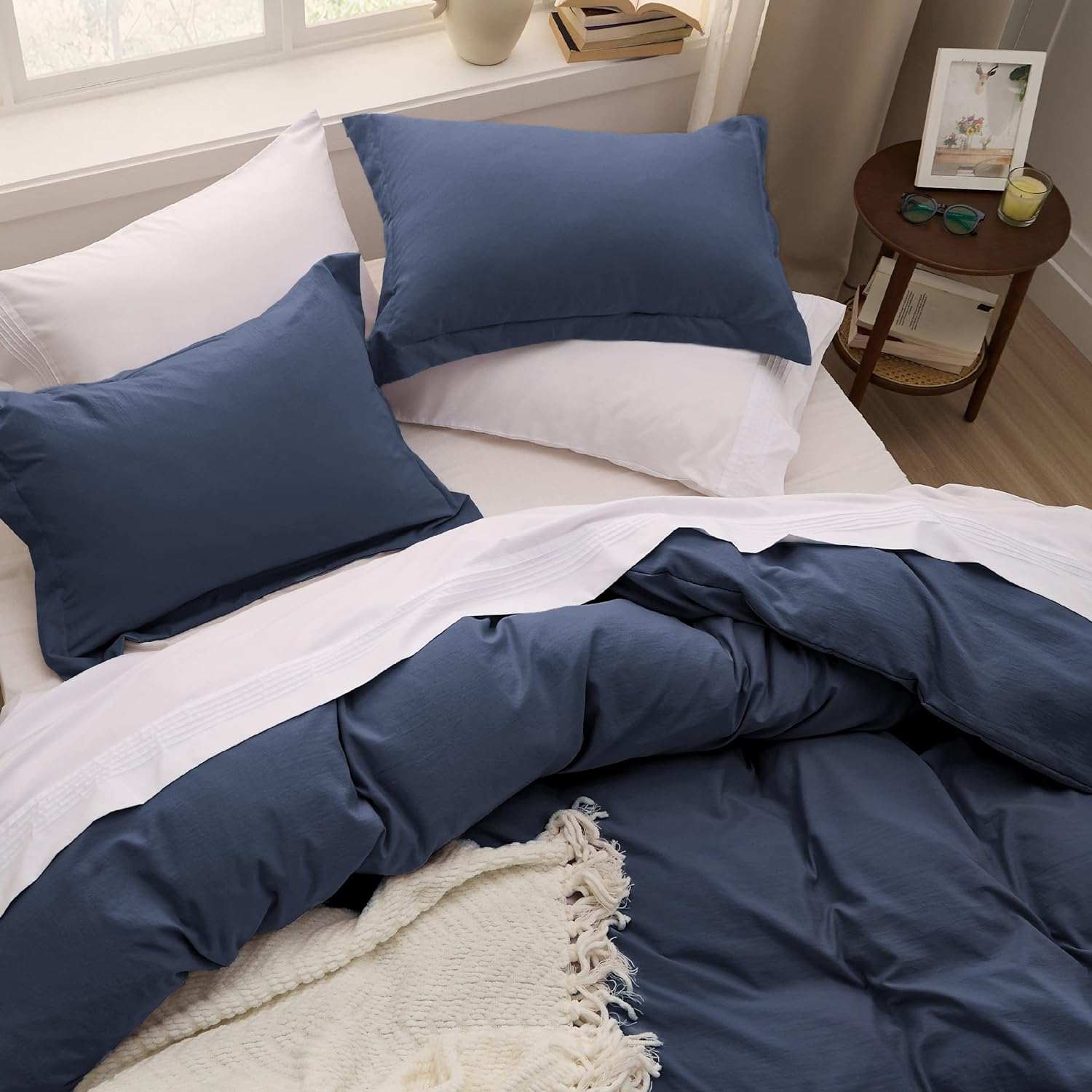 Overhead view of a neatly made bed featuring a navy blue bedding set. The set includes pillowcases and a comforter, paired with white sheets. A textured white throw blanket is placed at the foot of the bed. The bed is positioned near a large window with sheer curtains, allowing natural light to fill the room. A bedside table holds a book, glasses, a framed picture, and a candle, adding a cozy and inviting touch to the scene