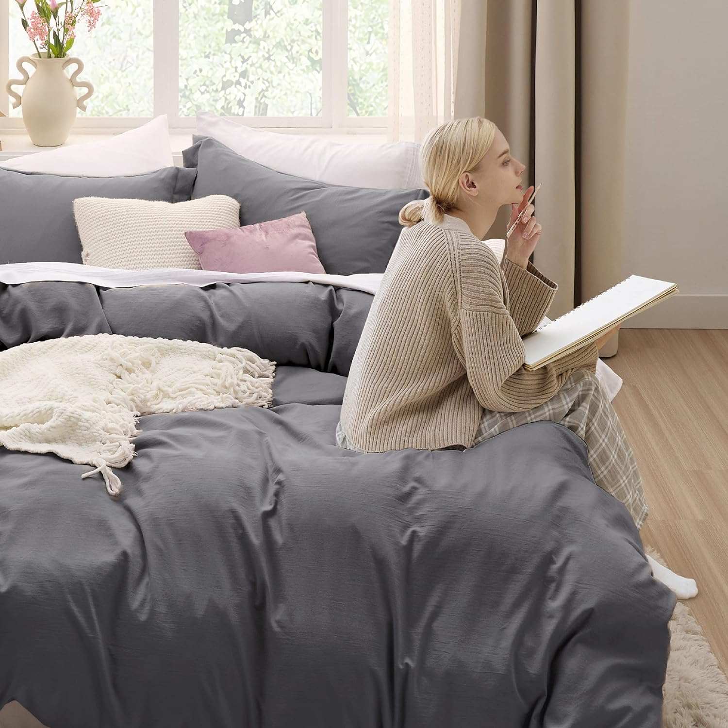 Cozy bedroom with charcoal bedding, woman sketching on bed, soft lighting, and decorative pillows. Comfortable and serene ambiance.