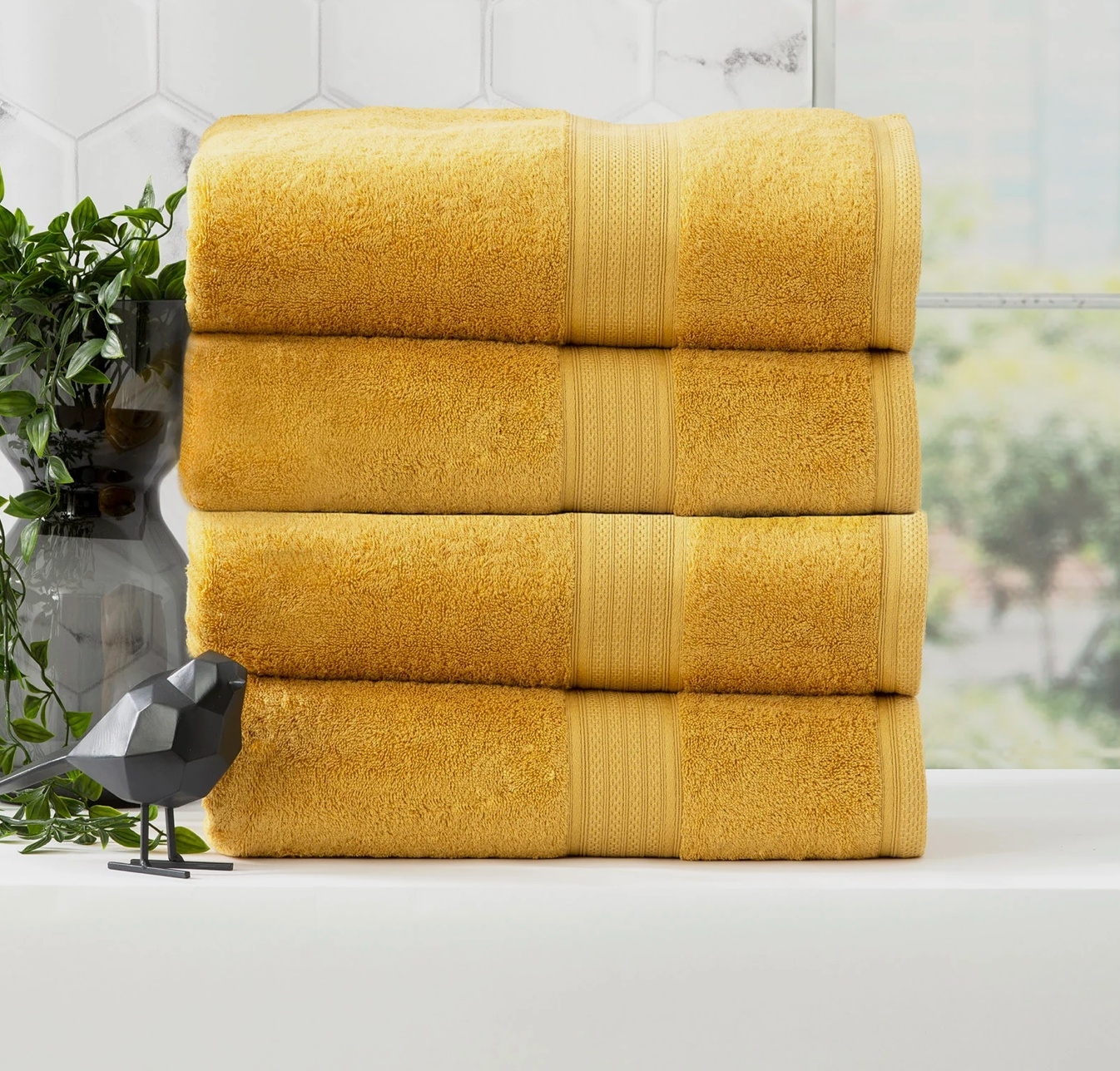 Four folded golden yellow Adorearth™ Organic Bamboo Towels stacked beside a decorative plant and black bird figurine.