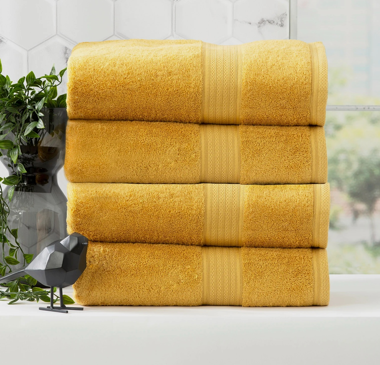 Four folded yellow Adorearth™ organic bamboo towels stacked on a white surface near a potted plant and bird figurine in a modern bathroom.